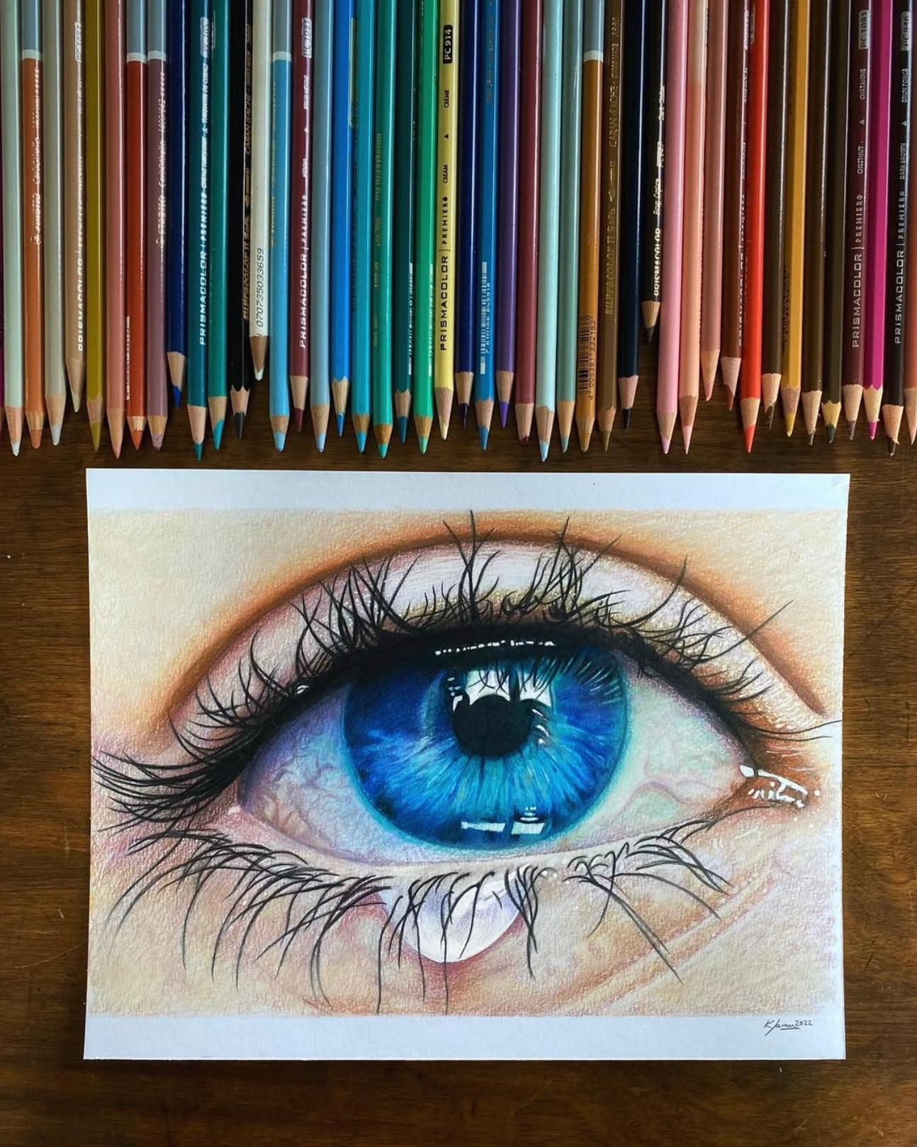 Detailed coloured-pencil drawing of a blue eye with pencils arranged above it.