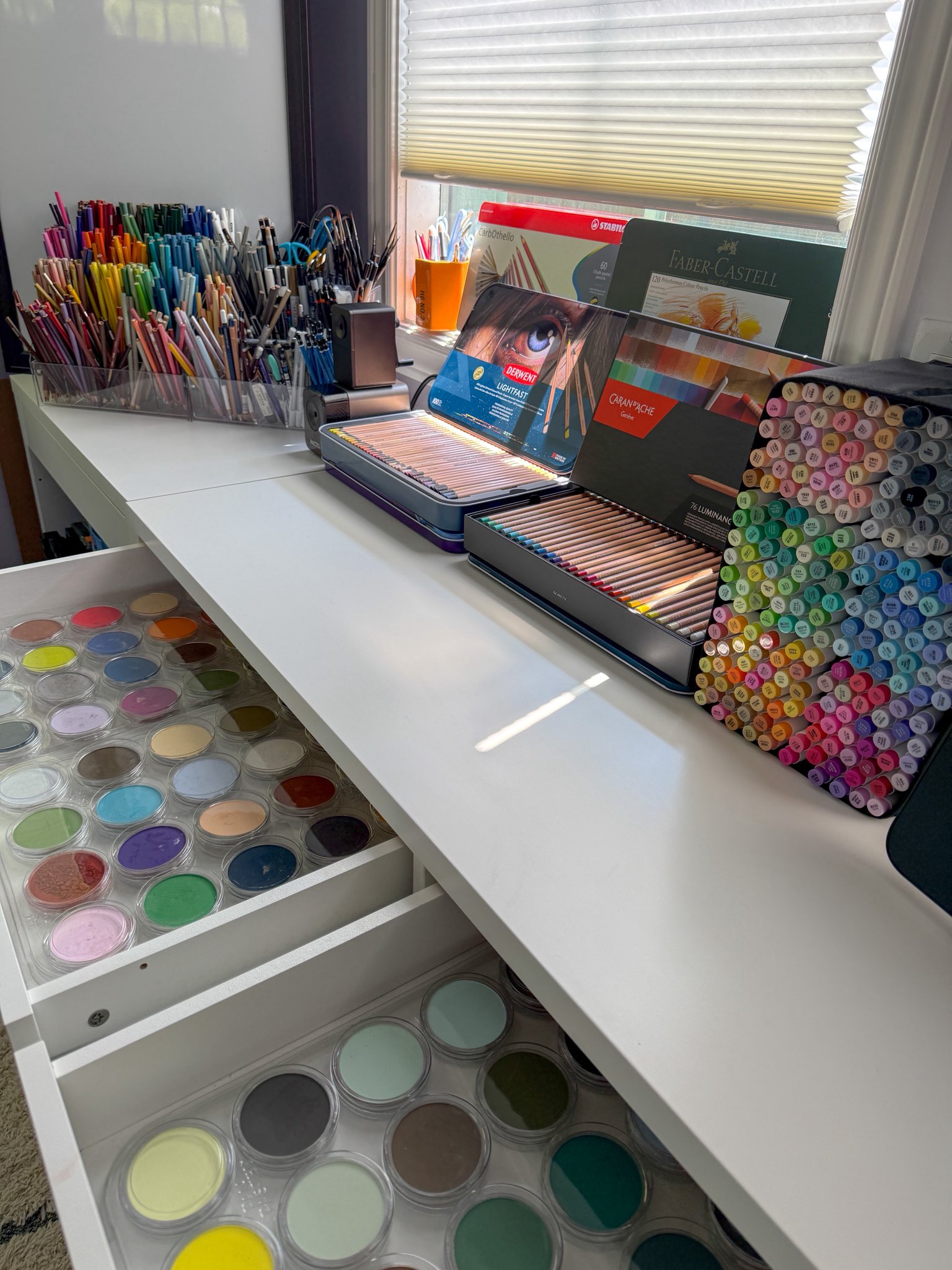 A sunlit desk filled with coloured pencils, markers and pastel pigments.
